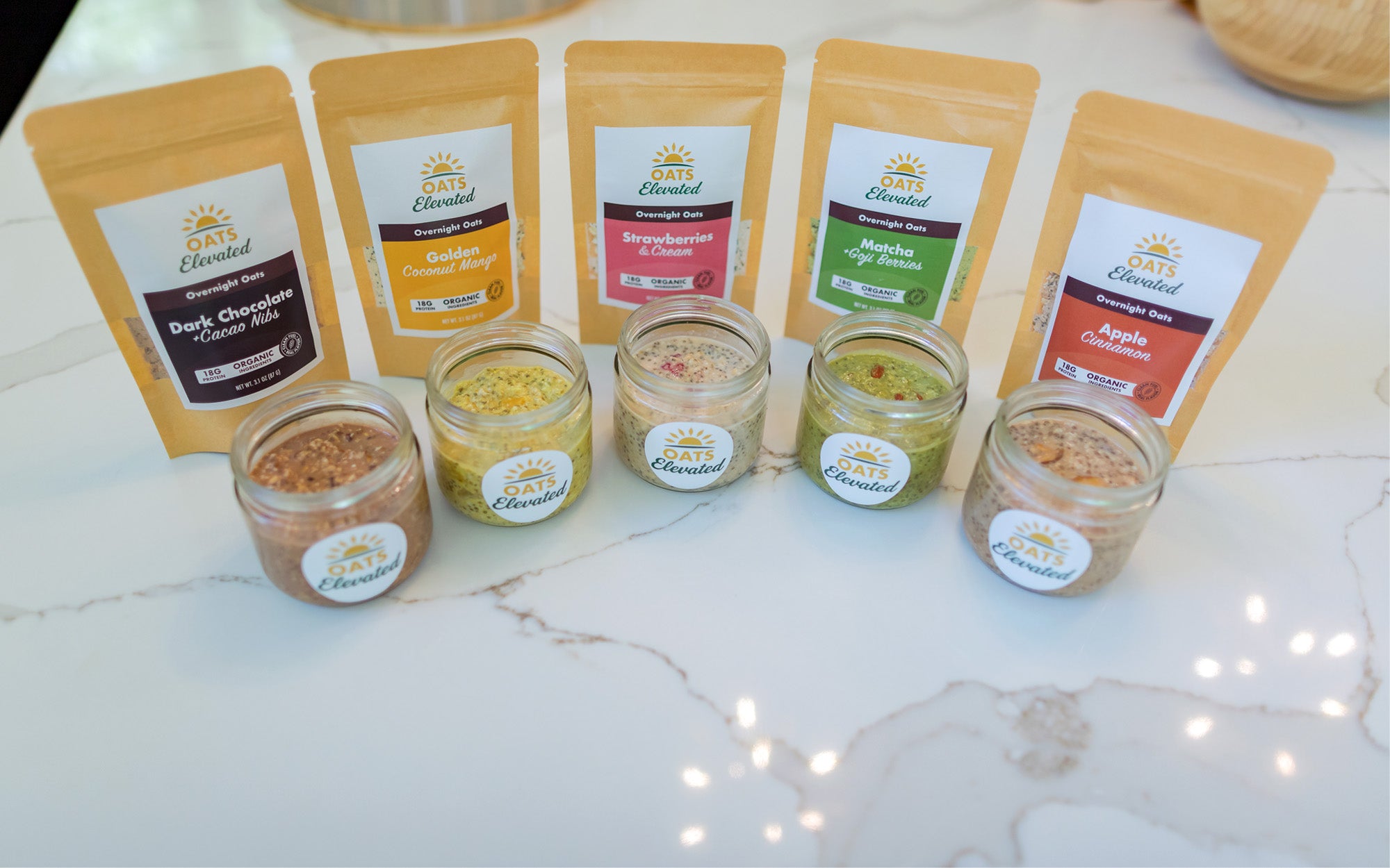 Five different package of Oats Elevated overnight oats standing behind a jar of prepared oats for each flavor.